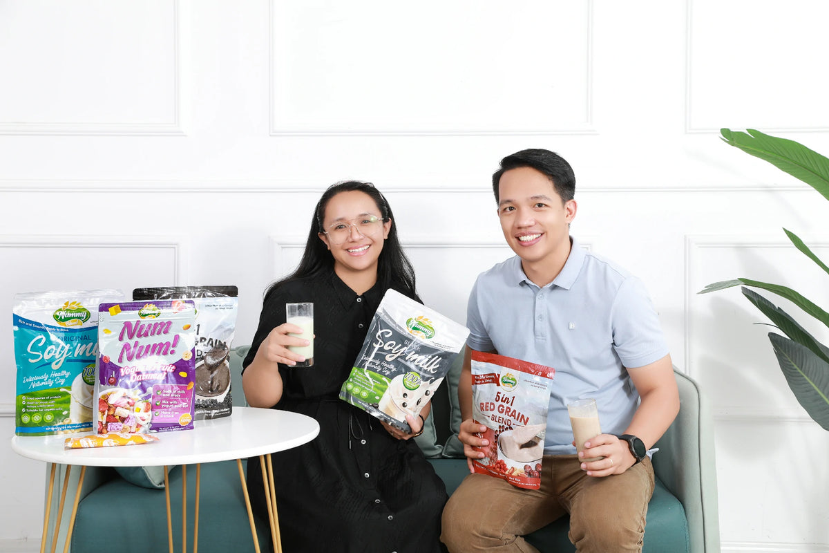 Two adults sitting together on a sofa, smiling and holding glasses of plant based drinks, with multiple Nummy Blend product packages displayed on a small table beside them in a bright, modern living space.